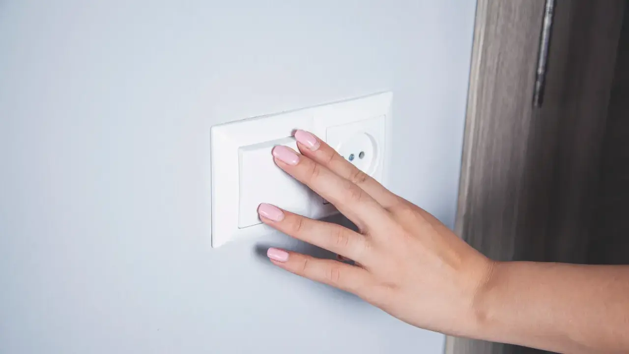 Main d'une femme aux ongles roses actionnant un interrupteur blanc, prêt pour le branchement d'une lampe de chevet.
