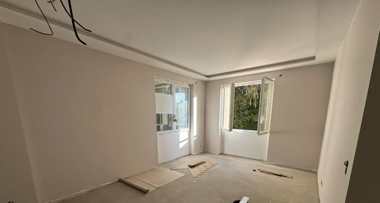 Chambre en cours de rénovation avec un faux plafond élégant, deux fenêtres ouvertes sur la verdure et des matériaux de construction au sol.
