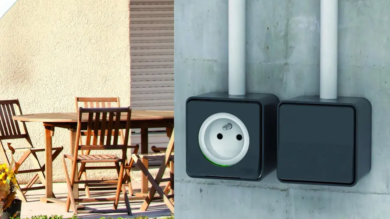 Branchement prise extérieur sur mur de béton. Une prise électrique noire est visible, avec une autre à côté. Une table et des chaises en bois sont sur une terrasse.