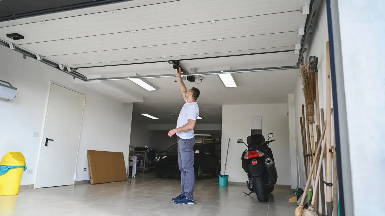 Un homme ajuste le moteur de la porte de garage électrique. Un scooter et des outils sont garés à proximité.