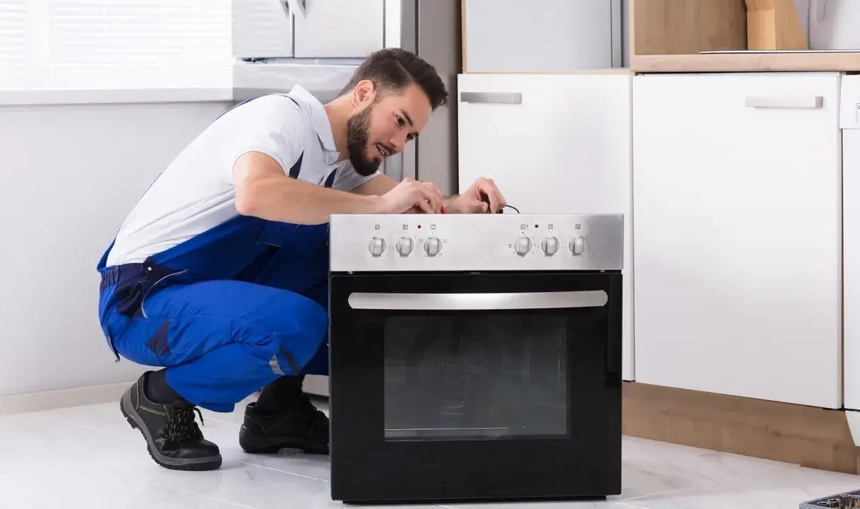 Un technicien en salopette bleue répare une prise four. Il est accroupi devant l'appareil, concentré sur les fils.