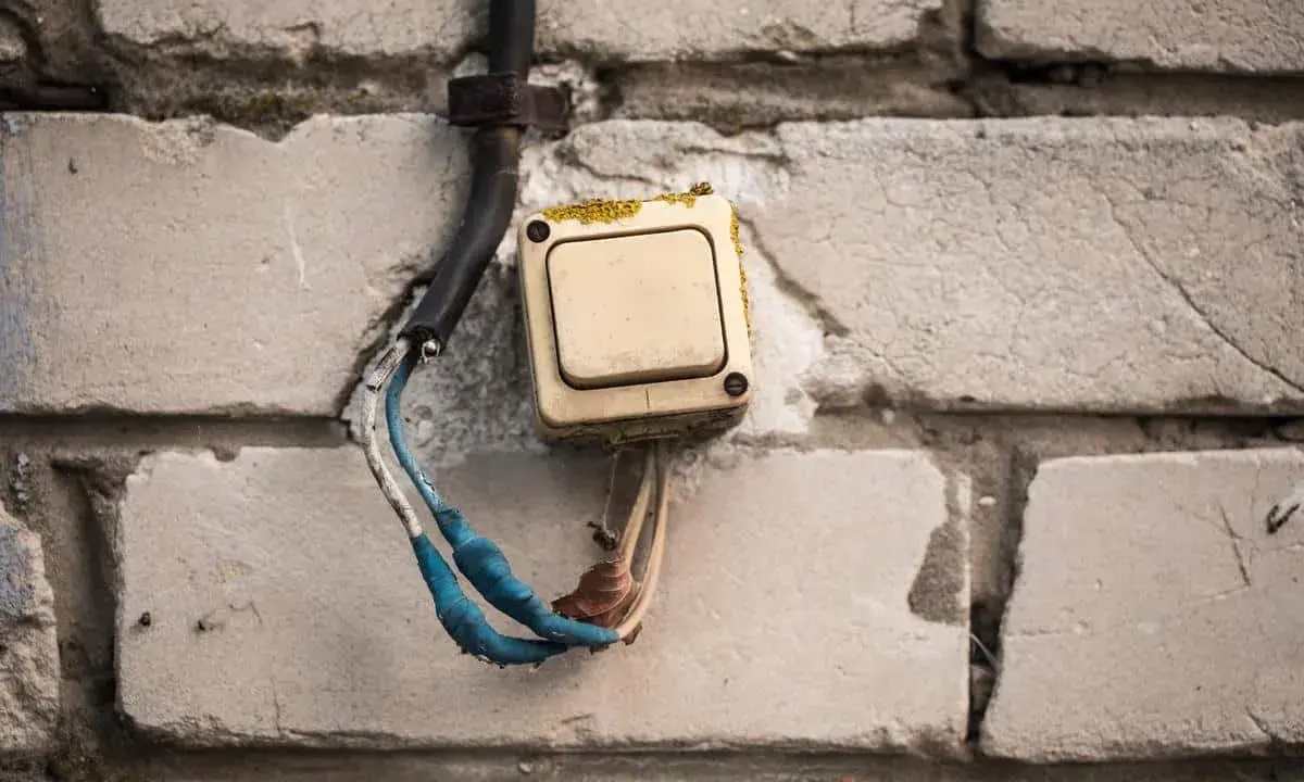 Interrupteur électrique beige en saillie sur un mur de briques, fils dénudés bleus et blancs.