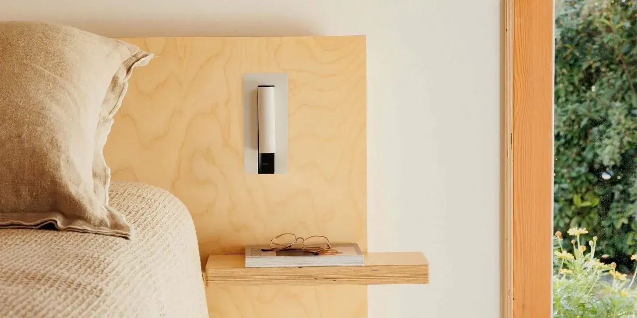 Applique murale au design épuré, à la bonne hauteur pour une tête de lit en bois clair. Lunettes posées sur un livre.