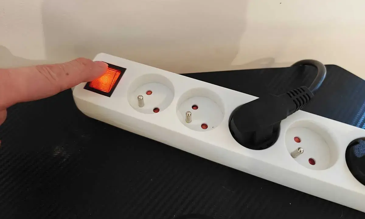 Doigt appuyant sur l'interrupteur d'une multiprise blanche, plusieurs prises vides et une prise avec un câble noir.