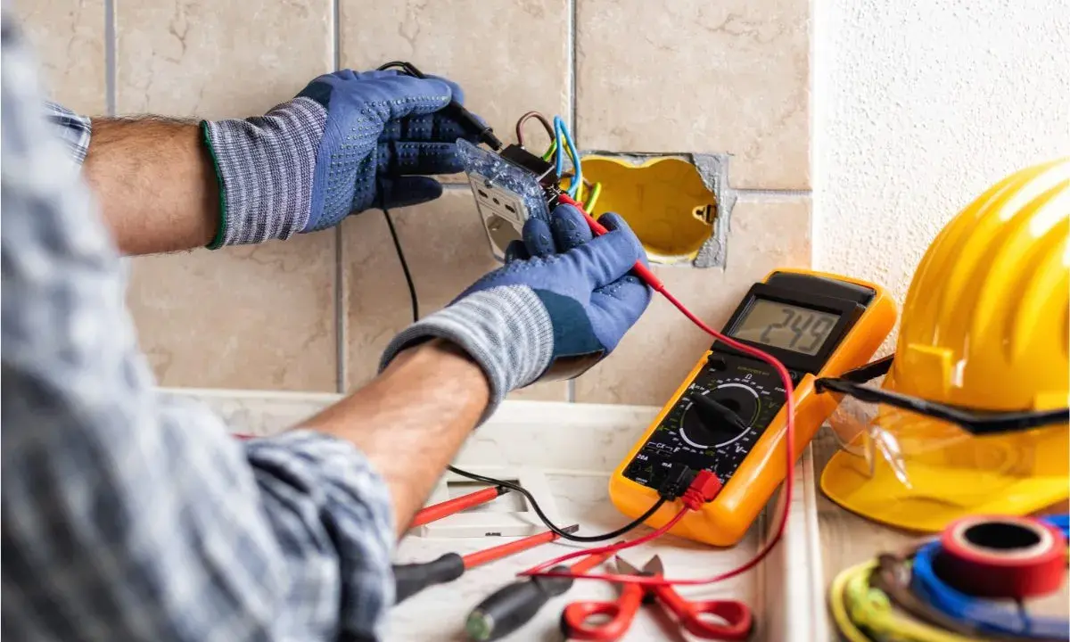 Un électricien vérifie un circuit électrique avec un multimètre. Des outils et un casque de sécurité sont visibles.