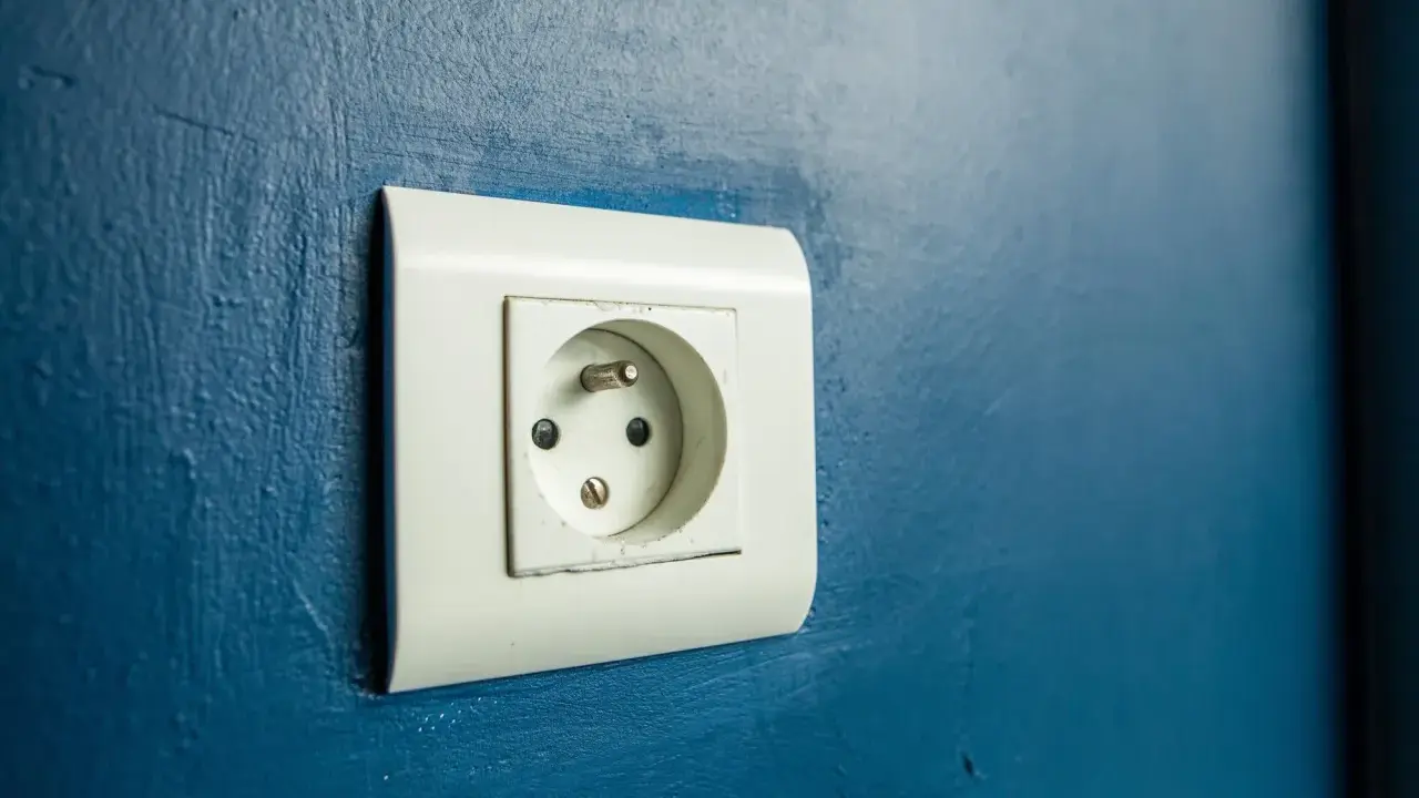 Prise électrique murale blanche sur fond bleu. Le branchement prise de terre est visible, prêt à connecter un appareil.