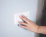 Main d'une femme aux ongles roses actionnant un interrupteur blanc, prêt pour le branchement d'une lampe de chevet.