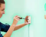 Une femme réalise un branchement de prise avec un tournevis. Un personnage de dessin animé, un ouvrier, fait un clin d'œil et un signe "OK".