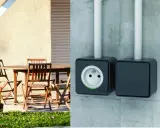 Branchement prise extérieur sur mur de béton. Une prise électrique noire est visible, avec une autre à côté. Une table et des chaises en bois sont sur une terrasse.