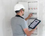 Un technicien en casque blanc consulte un schéma électrique sur sa tablette, devant un tableau de disjoncteurs. Il supervise l'installation électrique d'un nouveau gtl logement.
