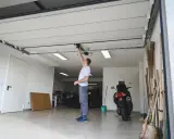 Un homme ajuste le moteur de la porte de garage électrique. Un scooter et des outils sont garés à proximité.