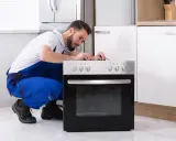 Un technicien en salopette bleue répare une prise four. Il est accroupi devant l'appareil, concentré sur les fils.