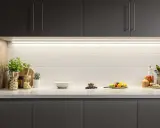 Éclairage subtil sous les armoires sombres, mettant en valeur le carrelage blanc et les objets décoratifs. Une **reglette LED design** illumine la cuisine.