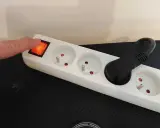 Doigt appuyant sur l'interrupteur d'une multiprise blanche, plusieurs prises vides et une prise avec un câble noir.