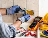 Un électricien vérifie un circuit électrique avec un multimètre. Des outils et un casque de sécurité sont visibles.