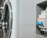 Disjoncteur machine à laver, prêt à protéger l'appareil. L'image montre un disjoncteur blanc et bleu, avec des fils orange et jaunes connectés, à côté d'une machine à laver.
