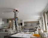 Un homme sur une échelle installe un spot dimmable au plafond d'un salon moderne.