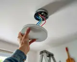 Installation d'un plafonnier : une main tient le cache, révélant le kit de fixation et les fils électriques colorés.