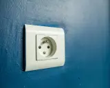 Prise électrique murale blanche sur fond bleu. Le branchement prise de terre est visible, prêt à connecter un appareil.