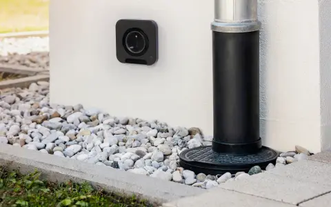 Une prise femelle étanche noire est fixée au mur blanc, à côté d'une descente d'eau.