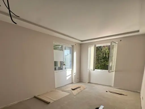 Chambre en cours de rénovation avec un faux plafond élégant, deux fenêtres ouvertes sur la verdure et des matériaux de construction au sol.
