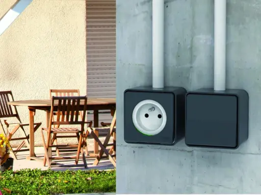 Branchement prise extérieur sur mur de béton. Une prise électrique noire est visible, avec une autre à côté. Une table et des chaises en bois sont sur une terrasse.