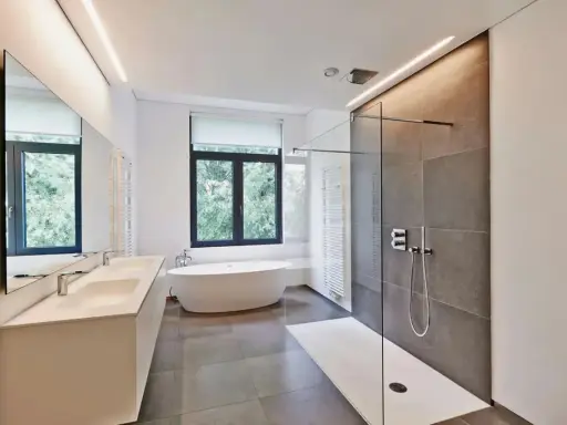 Salle de bain moderne avec baignoire îlot, double vasque et douche à l'italienne. L'éclairage intégré en hauteur applique une ambiance douce.
