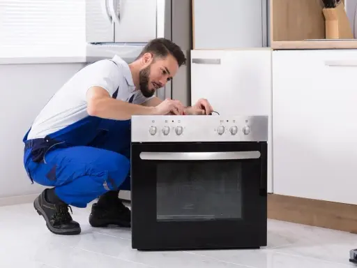 Un technicien en salopette bleue répare une prise four. Il est accroupi devant l'appareil, concentré sur les fils.