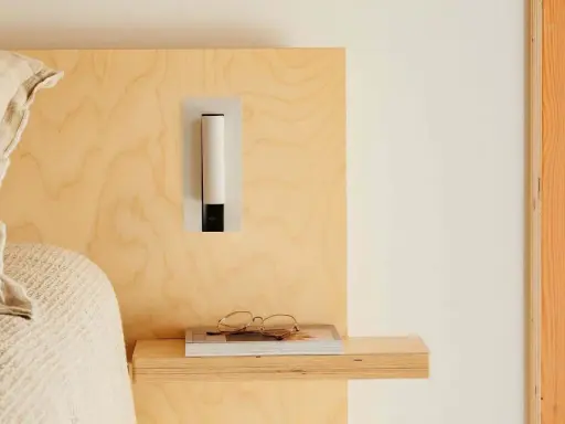 Applique murale au design épuré, à la bonne hauteur pour une tête de lit en bois clair. Lunettes posées sur un livre.