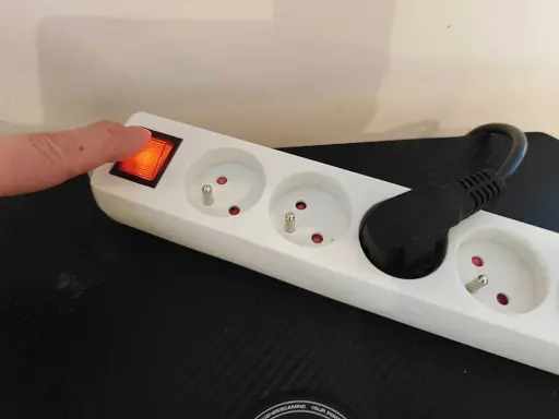 Doigt appuyant sur l'interrupteur d'une multiprise blanche, plusieurs prises vides et une prise avec un câble noir.