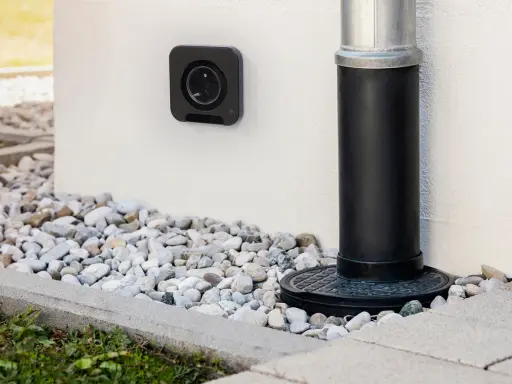 Une prise femelle étanche noire est fixée au mur blanc, à côté d'une descente d'eau.