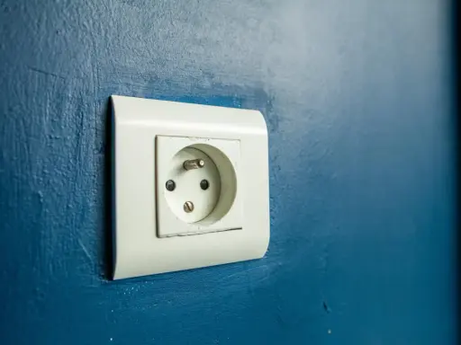 Prise électrique murale blanche sur fond bleu. Le branchement prise de terre est visible, prêt à connecter un appareil.