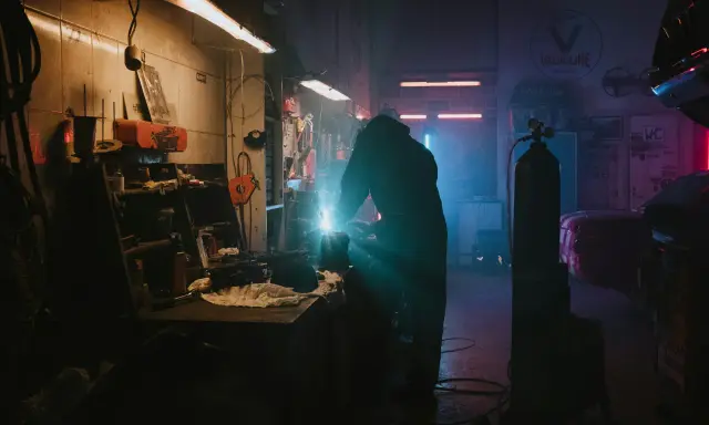 Un soudeur travaille dans un garage sombre, éclairé par des néons. La question "dans quel sens installer un néon dans un garage" plane dans l'air.