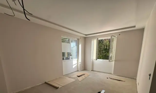 Chambre en cours de rénovation avec un faux plafond élégant, deux fenêtres ouvertes sur la verdure et des matériaux de construction au sol.