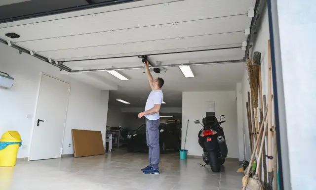 Un homme ajuste le moteur de la porte de garage électrique. Un scooter et des outils sont garés à proximité.