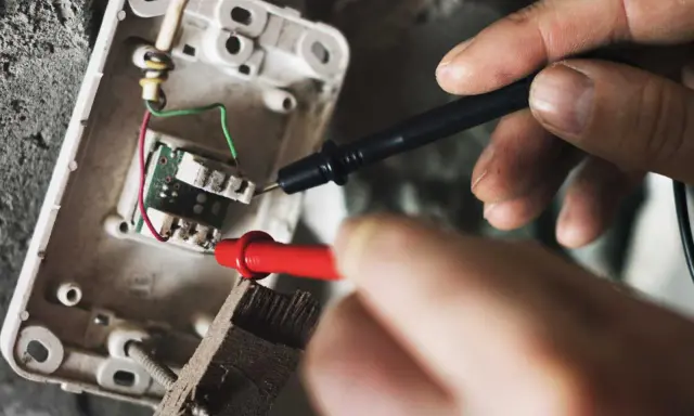 Mains électriques sous tension, un technicien vérifie un fusible qui chauffe avec un multimètre.