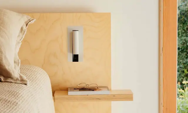 Applique murale au design épuré, à la bonne hauteur pour une tête de lit en bois clair. Lunettes posées sur un livre.