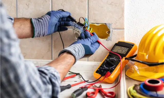 Un électricien vérifie un circuit électrique avec un multimètre. Des outils et un casque de sécurité sont visibles.