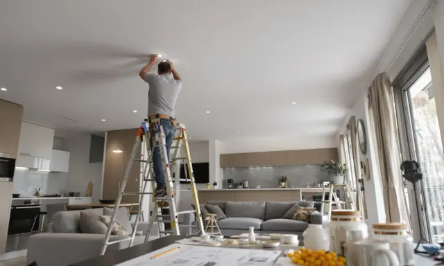 Un homme sur une échelle installe un spot dimmable au plafond d'un salon moderne.