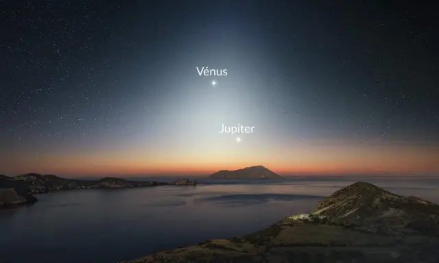 Le ciel étoilé, baigné d'une douce **lumière du jour** naissante, révèle Vénus et Jupiter au-dessus d'une côte rocheuse et d'une mer calme.