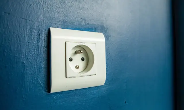 Prise électrique murale blanche sur fond bleu. Le branchement prise de terre est visible, prêt à connecter un appareil.