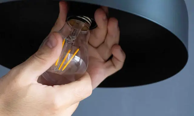 Mains tenant une ampoule LED à filament, prête à être installée. La chaleur de l'ampoule est symbolisée par ses filaments jaunes lumineux.