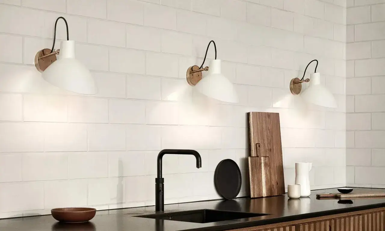 Trois appliques murales blanches avec un bras noir et une base dorée, éclairant un mur de carreaux blancs. Le branchement applique murale avec interrupteur est discret.