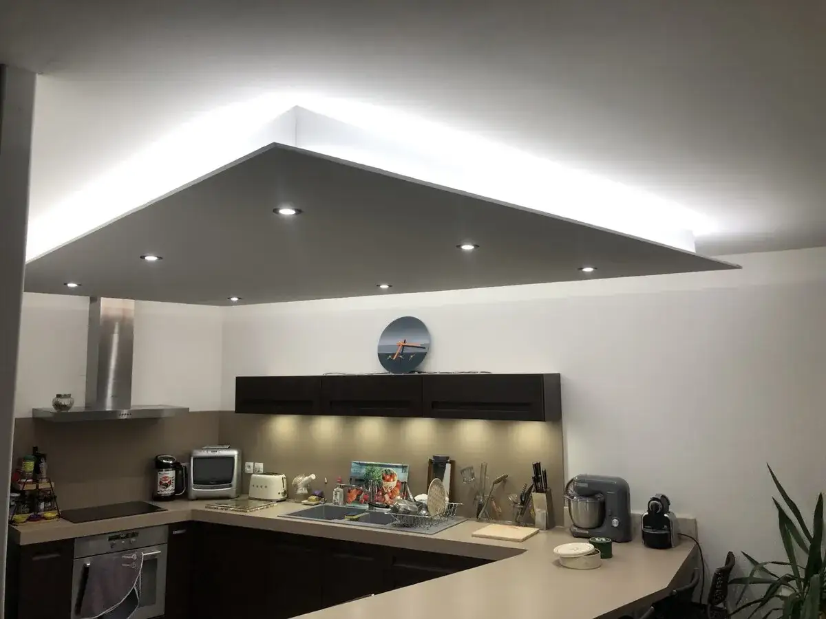 Cuisine moderne avec un faux plafond illumin&eacute;, spots encastr&eacute;s et &eacute;clairage indirect. Un plan de travail en L avec &eacute;vier et divers appareils &eacute;lectrom&eacute;nagers.