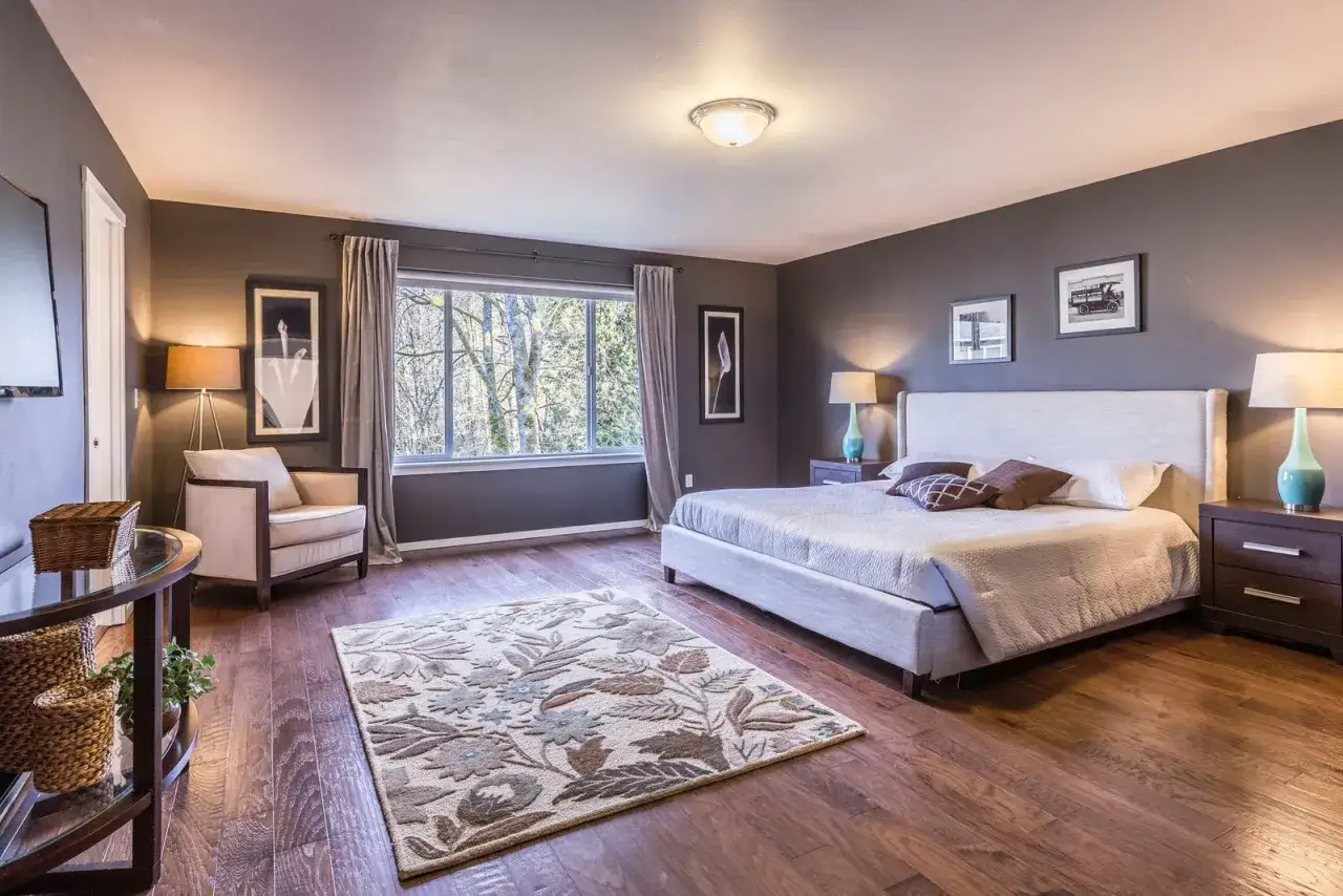 Chambre douillette avec lit king-size, tapis fleuri et fauteuil. L'&eacute;clairage doux cr&eacute;e une ambiance couleur blanc chaud.