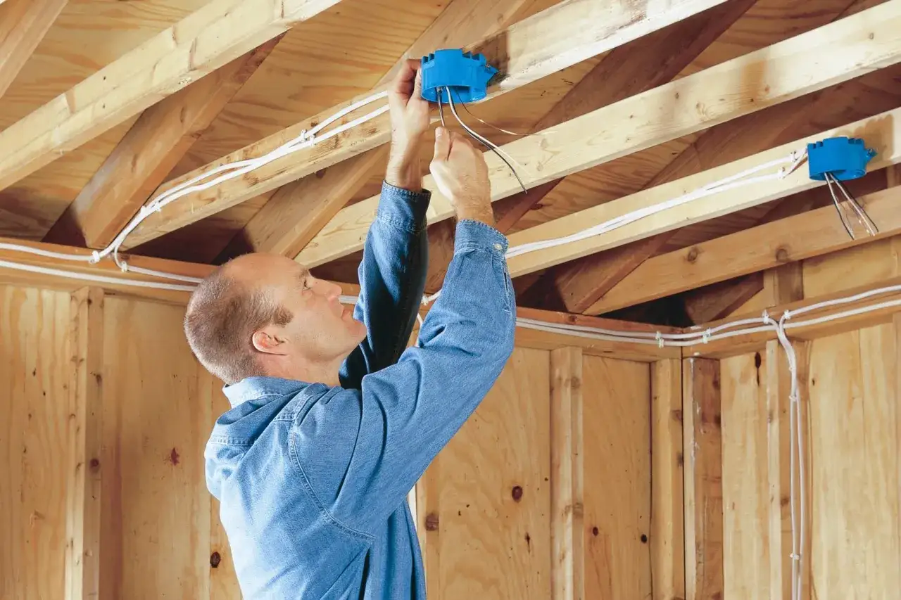Un homme installe des bo&icirc;tiers &eacute;lectriques bleus dans le plafond en bois d'un garage, pr&eacute;parant l'&eacute;lectricit&eacute;.
