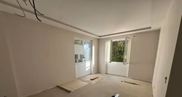 Chambre en cours de rénovation avec un faux plafond élégant, deux fenêtres ouvertes sur la verdure et des matériaux de construction au sol.
